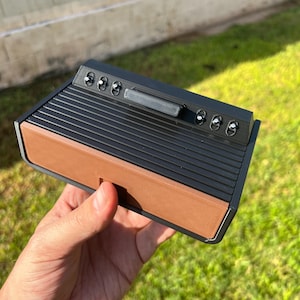 May include: A black and brown retro-style game console with several buttons and a handle. The console has a ribbed top and a brown base. The device is held in a hand against a background of green grass.