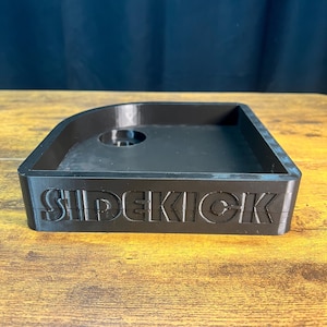 May include: A black 3D-printed tray with the word "SIDEKICK" embossed on the front. The tray has a rectangular shape with rounded corners and a small circular indentation in the center. The tray is sitting on a wooden surface.