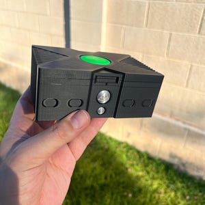 May include: A black, 3D-printed Xbox console replica. The model features a green power button, silver details, and a textured surface. The console is held against a brick wall and green grass.