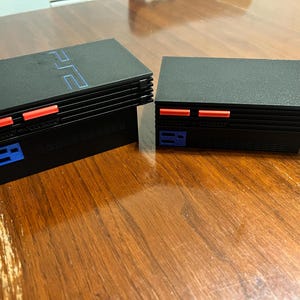 May include: Two black PlayStation 2 consoles. The top of each console has the PlayStation 2 logo in blue. Each console has red and blue accents on the front.