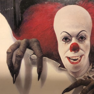 May include: A close-up image of a clown with white face paint, red nose, and red hair. The clown has dark eye makeup and a sinister smile. A clawed hand reaches out from the side, adding to the unsettling effect. The image evokes a horror theme.