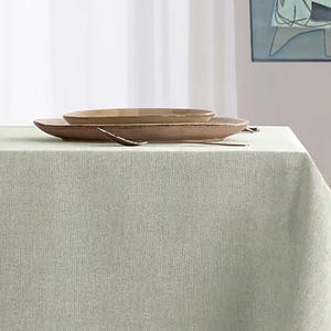May include: A light green tablecloth with two brown ceramic plates and a spoon. The plates are stacked on top of each other. The background includes a white curtain and a painting.