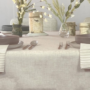 May include: A dining table setting with a light beige linen tablecloth. The table is set with brown bowls, plates, silverware, and striped napkins. Decorative birch bark vases and clear glass vases hold floral arrangements.