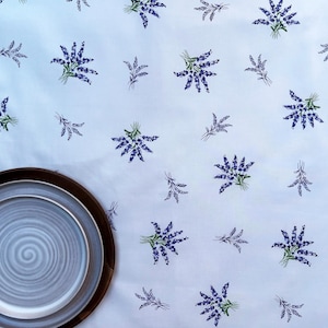May include: A white tablecloth featuring a repeating pattern of small purple lavender sprigs with green stems, paired with a round plate that has a light blue center and a dark brown rim, creating a simple and clean table setting arrangement.