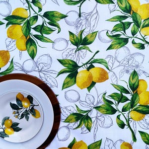 May include: A white tablecloth with a vibrant lemon and leaf pattern. The design features yellow lemons and green leaves, with a background of sketched lemon outlines. A wooden charger and two white plates are in the foreground, one with a lemon branch design.