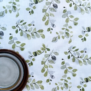 May include: A white tablecloth with a watercolor pattern of green and gray leaves and berries. A brown plate with a gray plate on top is in the lower left corner. The tablecloth is likely linen.