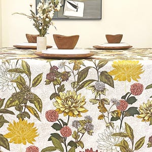 May include: A rectangular tablecloth featuring a botanical print of yellow chrysanthemums, pink flower clusters, and green foliage on a white background. The table is set with wooden bowls, white plates, and a vase of dried flowers.