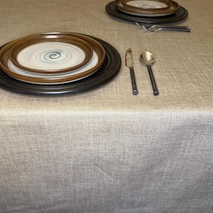May include: A beige textured tablecloth featuring two place settings. Each setting includes a large black charger plate, a brown-rimmed dinner plate with a blue spiral center, a silver knife, a silver spoon, and two silver forks.