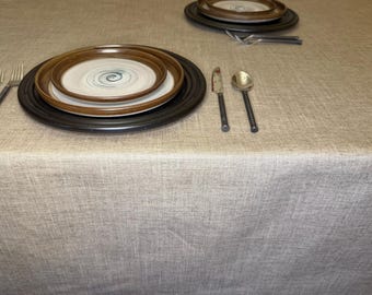 May include: A beige textured tablecloth featuring two place settings. Each setting includes a large black charger plate, a brown-rimmed dinner plate with a blue spiral center, a silver knife, a silver spoon, and two silver forks.