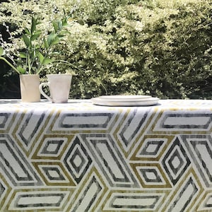May include: A table setting with a geometric patterned tablecloth in shades of gray, white, and yellow. A vase with greenery, a mug, and a plate are on the table. The background features lush green foliage.