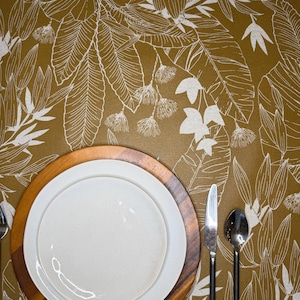 May include: A table setting featuring a white ceramic plate centered on a round wooden charger, placed on a mustard yellow tablecloth with a white botanical leaf pattern. A stainless steel knife and spoon are positioned to the right.
