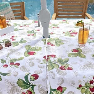 May include: A white tablecloth featuring a repeating pattern of red strawberries and green leaves. The fabric has a central opening for a patio umbrella pole. The design includes detailed illustrations of ripe berries and foliage.