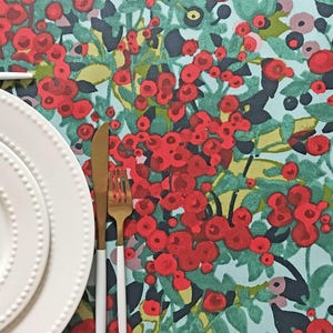 May include: A table setting featuring two white plates with beaded edges, a gold-toned knife and fork with white handles, and a tablecloth patterned with red berries and green foliage on a light blue background.