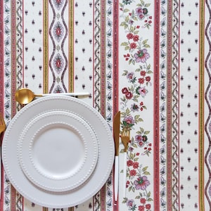 May include: A white tablecloth with a repeating vertical pattern of floral vines in pink and green, alongside decorative geometric borders. A set of two stacked white plates with beaded rims is placed with gold-toned cutlery featuring white handles.