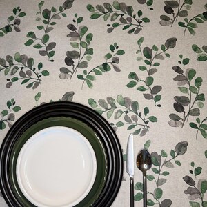 May include: A table setting featuring a white plate nested inside a dark green plate and a black charger plate. A silver knife and spoon are placed to the right. The items rest on a light-colored fabric tablecloth with a green and gray leaf pattern.