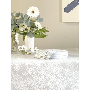 May include: A dining table setting with white plates, a striped napkin, and a vase of white flowers. The tablecloth has a white coral pattern. A second vase with flowers is visible.