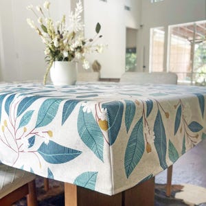 May include: A rectangular tablecloth featuring a botanical print with teal and blue leaves, small white flowers, and mustard yellow buds on a light beige background. The fabric is draped over a table, showcasing a repeating pattern of foliage.