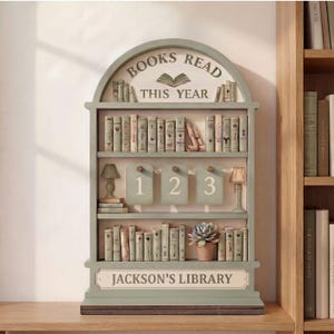 May include: A decorative bookshelf with the text "BOOKS READ THIS YEAR" and "JACKSON'S LIBRARY". The bookshelf is painted in a muted green color and features miniature books, lamps, and numbered tags. A small potted succulent is also visible.