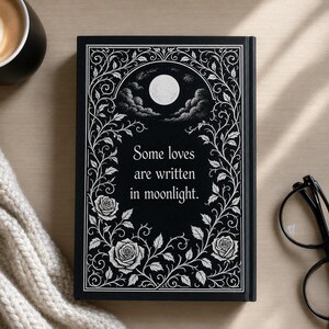 Może przedstawiać: Czarny notatnik z cytatem "Some loves are written in moonlight." Okładka przedstawia biały kwiatowy wz&oacute;r z r&oacute;żami, pełnią księżyca i chmurami. Notatnik leży na drewnianej powierzchni, z filiżanką kawy i okularami w pobliżu.