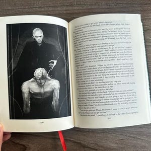 Może przedstawiać: Otwarta książka z czerwoną wstążką jako zakładką, z czarno-białą ilustracją dw&oacute;ch postaci. Ilustracja przedstawia mroczną postać g&oacute;rującą nad siedzącą osobą. Tekst jest widoczny na stronach.