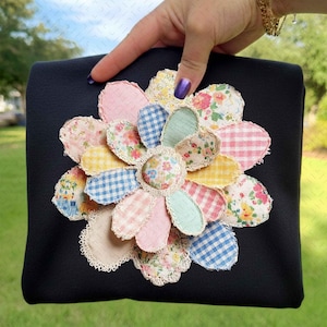 May include: A black square clutch bag with a large, colourful flower appliqué. The flower features pastel pink, yellow, blue, and floral patterned fabric petals. The centre of the flower is a small, round fabric piece with a floral pattern. The edges have a cream-coloured lace trim.