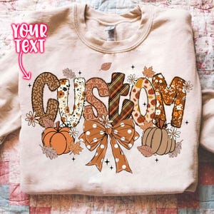 Pode incluir: Camisola bege com a palavra "CUSTOM" num design com tema de outono. As letras apresentam padrões como estampado de leopardo, xadrez e desenhos florais, com abóboras, folhas e um laço. O texto "YOUR TEXT" está acima.