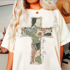 Könnte beinhalten: Cremefarbenes T-Shirt mit einem großen Kreuzdesign aus Camouflage-Stoff mit ausgefransten Kanten. Das Kreuz ist in Grün-, Braun- und Grautönen gehalten. Die Worte "He is Risen" sind vertikal rechts vom Kreuz in schwarzer Schrift geschrieben.
