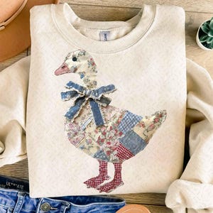 Könnte beinhalten: Cremefarbenes Sweatshirt mit einem Gänsemotiv aus Patchwork. Die Gans hat einen floral gemusterten Kopf und Körper, eine blaue Schleife und rot-weiß karierte Füße. Das Sweatshirt hat ein strukturiertes Muster und einen Rundhalsausschnitt.