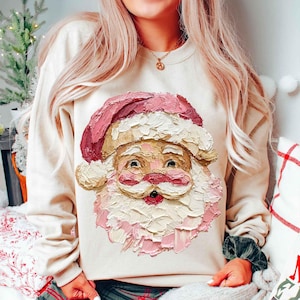 Könnte beinhalten: Ein cremefarbenes Sweatshirt mit einem strukturierten Weihnachtsmann-Gesicht in Rosa-, Weiß- und Beigetönen. Das Weihnachtsmann-Design hat einen rosa Hut und Bart, mit rosiger Nase und Wangen. Das Sweatshirt ist ein festliches Kleidungsstück für die Feiertage.