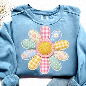 May include: A light blue sweatshirt featuring a large, appliqued flower design. The flower has a yellow center and petals in various pastel patterns, including polka dots, gingham, and floral prints. The sweatshirt has a crew neck and long sleeves.