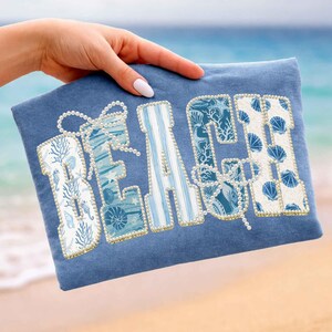 May include: A blue t-shirt with the word "BEACH" in large, decorative letters. The letters are embellished with pearls and feature various beach-themed patterns, including seashells and coral. The background is a blurred beach scene.