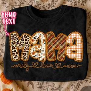 Puede incluir: Sudadera negra con la palabra "MAMA" en letras grandes y decorativas. Las letras están estampadas con diseños de leopardo, flores, cuadros y cuadros vichy. Debajo, los nombres "emily", "liam" y "anna".