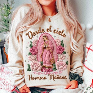 May include: Beige sweatshirt featuring an embroidered design of a religious figure in pink, surrounded by pink roses and the text "Desde el cielo Una Hermosa Mañana" in brown. The design is centered on the front of the sweatshirt.