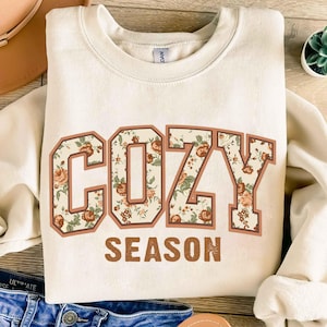 Può includere: Felpa color crema con le parole "COZY SEASON" in un design floreale. La parola "COZY" è in uno stile a blocchi con un motivo floreale, e "SEASON" è in un semplice font marrone. La felpa ha uno scollo tondo.
