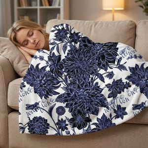 Puede incluir: Una manta blanca con un gran estampado floral en tonos azules y negros. El diseño presenta dalias y hojas estilizadas, con la palabra "flower" escrita en cursiva. La manta está sobre un sofá beige.