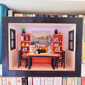 May include: A miniature diorama of a dining room scene, framed in black. The room features orange shelves, a table, chairs, and various miniature food items. The diorama is set against a backdrop of a landscape painting and is surrounded by books.