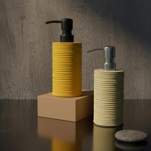 Può includere: Due dispenser di sapone a coste, uno giallo con una pompa nera e l'altro color crema con una pompa argentata, sono appoggiati su una superficie scura. Il dispenser giallo è su una piccola scatola beige. Due piccole pietre grigie sono in primo piano.