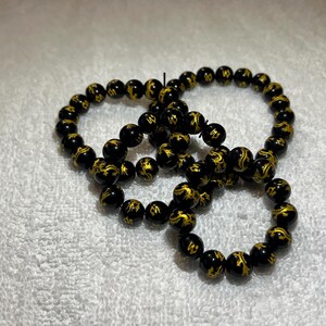 May include: Three black beaded bracelets with gold-colored dragon designs. The bracelets are made of round beads and are arranged in a circular pattern. The bracelets are on a white surface.