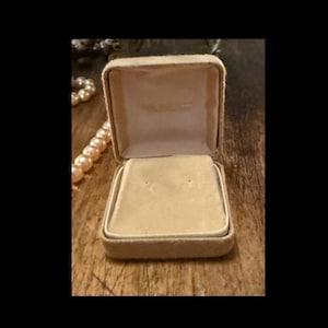 May include: An open, beige jewelry box with a soft, velvety interior. The box is square with a hinged lid and a small, rectangular space for a necklace or bracelet. A strand of pearls is visible in the background.
