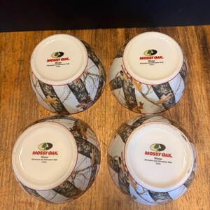 May include: Four ceramic bowls with a camouflage design in brown, white, and grey. The bowls have a white interior and a red rim. The text "MOSSY OAK. Winter" is printed on the base of each bowl.