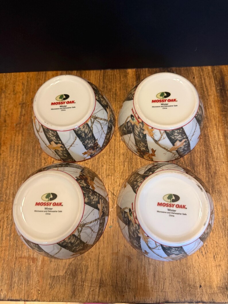 May include: Four ceramic bowls with a camouflage design in brown, white, and grey. The bowls have a white interior and a red rim. The text "MOSSY OAK. Winter" is printed on the base of each bowl.