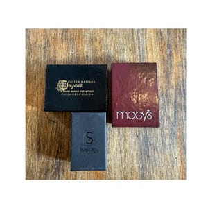 May include: Three small boxes on a wooden surface. One black box reads "UNITED NATIONS Bazaar GIFTS FROM ROUND THE WORLD PHILADELPHIA, PA." A burgundy box reads "macy's." A dark gray box reads "S SASHA BELL JEWELRY."
