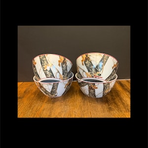 May include: Four white ceramic bowls with a camouflage pattern featuring brown, tan, and green elements. The bowls have a red rim and a dark interior. The bowls are stacked in pairs on a wooden surface.