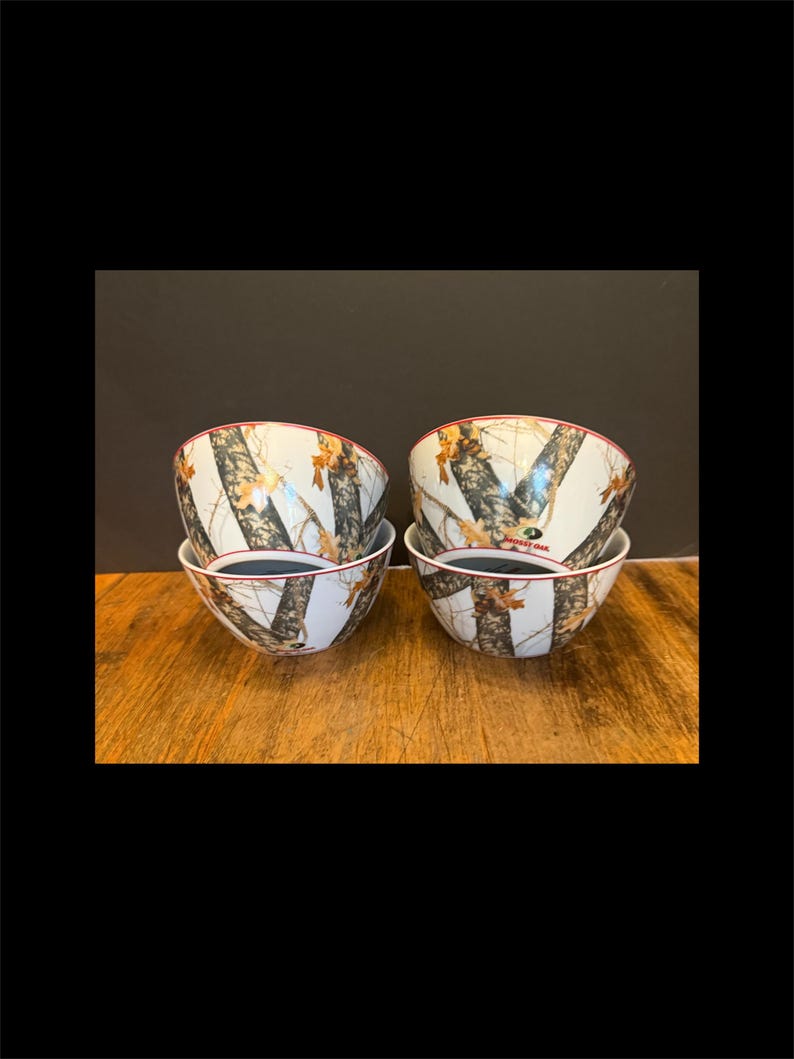 May include: Four white ceramic bowls with a camouflage pattern featuring brown, tan, and green elements. The bowls have a red rim and a dark interior. The bowls are stacked in pairs on a wooden surface.