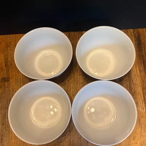 May include: Four white ceramic bowls arranged in a square. The bowls have a simple, rounded design with a smooth, glossy finish. The bowls are empty and appear to be new. The bowls are on a wooden surface.