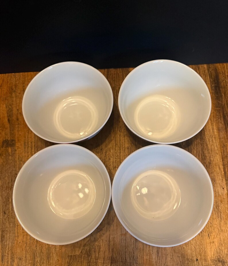 May include: Four white ceramic bowls arranged in a square. The bowls have a simple, rounded design with a smooth, glossy finish. The bowls are empty and appear to be new. The bowls are on a wooden surface.