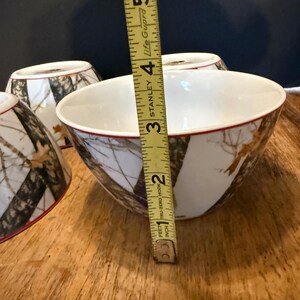 May include: White ceramic bowls with a camouflage pattern and a red rim. The bowls are approximately 10 cm in diameter. The bowls are on a wooden surface.