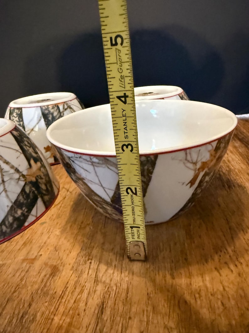 May include: White ceramic bowls with a camouflage pattern and a red rim. The bowls are approximately 10 cm in diameter. The bowls are on a wooden surface.