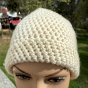 Könnte beinhalten: Eine cremefarbene, handgehäkelte Beanie-Mütze. Die Mütze hat ein strukturiertes, wabenartiges Stichmuster. Die Aufnahme ist eine Nahaufnahme, die die Details des Garns und der Häkelarbeit zeigt. Die Mütze ist ein Winteraccessoire.