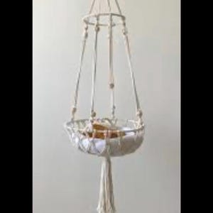 May include: A cream-colored macrame hanging basket featuring a circular frame, woven cord netting, and decorative wooden beads. The structure is suspended by multiple cords meeting at a top loop and terminates in a long, thick fringe tassel at the base.